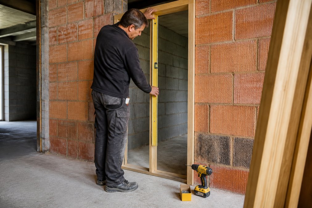 A joiner in work clothes holding a spirit level against an oak door frame set into a brick opening, with a cordless drill and screws resting on the floor nearby