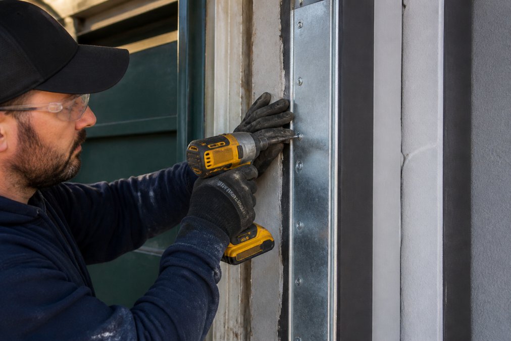 A maintenance worker in work gloves tightening screws on a galvanized steel jamb patch fitted over a repaired door frame section, bright workshop lighting
