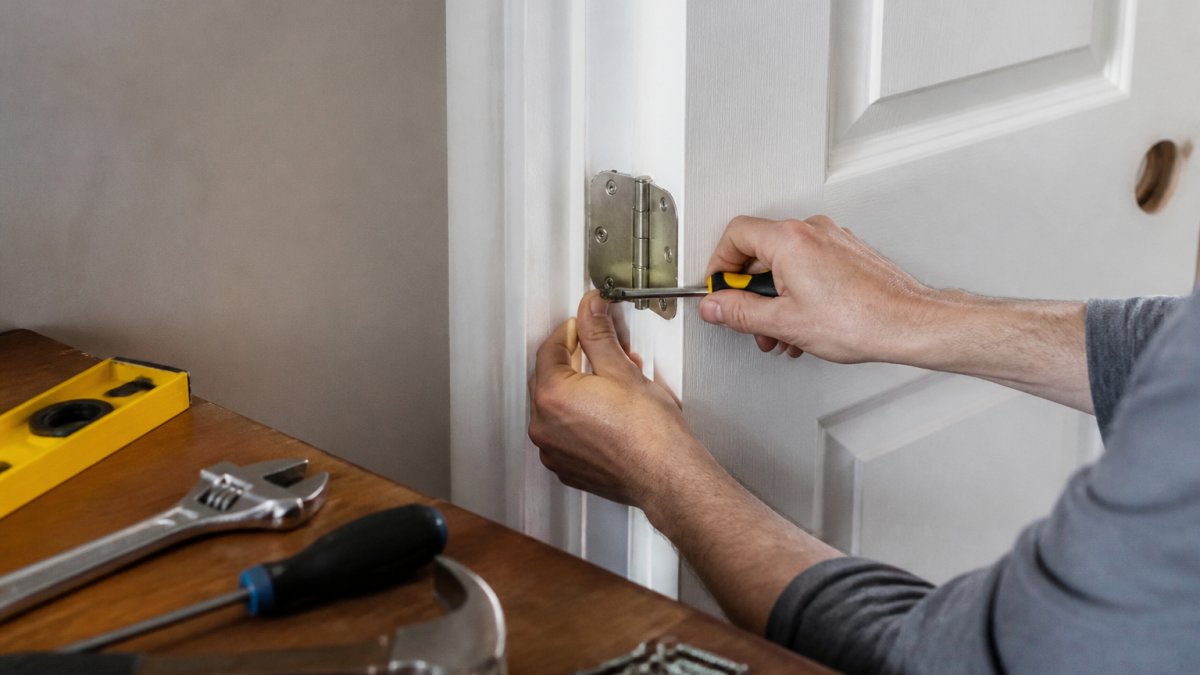 Homeowner's hands adjusting a door hinge with a screwdriver, basic tools including a level and adjustable wrench laid out on a wooden workbench