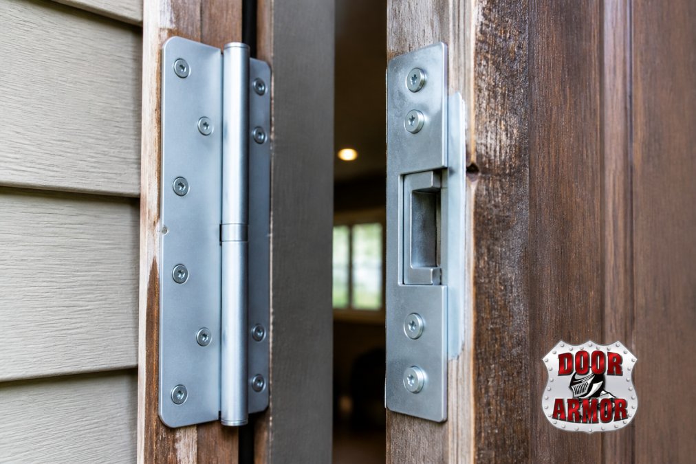 Close-up of a reinforced door frame with heavy-duty strike plate and hinge shields mounted on a residential entry door, showing galvanized steel hardware against weathered wood framing