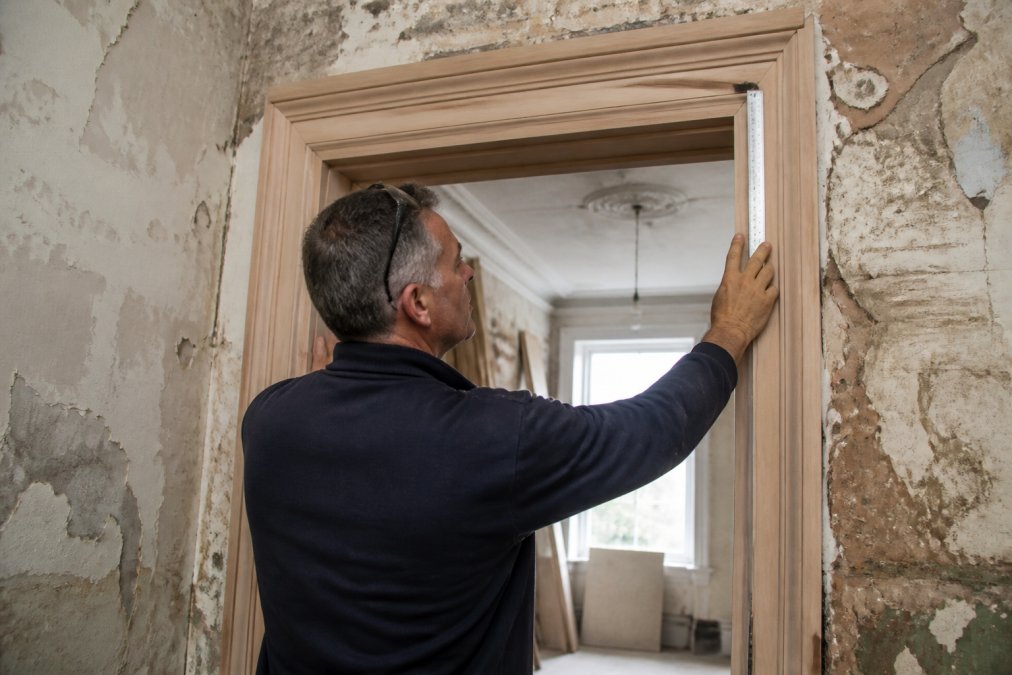 Craftsperson measuring and carefully fitting a custom wooden door frame into a period property opening, showing precise joinery work and attention to detail with natural workshop lighting