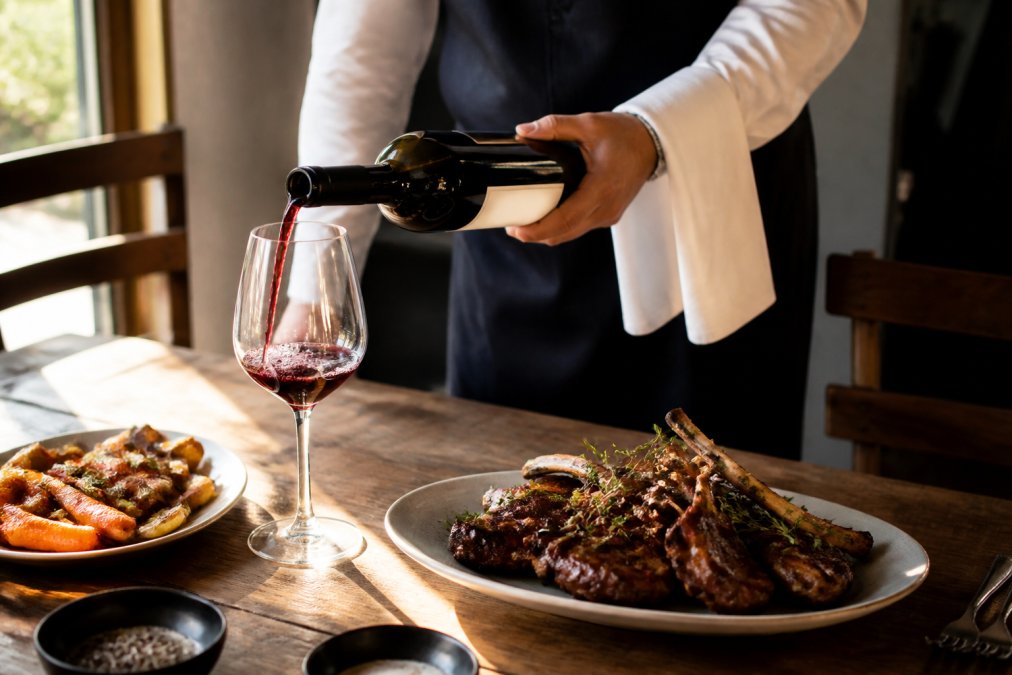 A sommelier pouring deep red Douro wine into a glass beside a plate of grilled lamb with herbs and roasted vegetables, natural afternoon light streaming across a wooden table
