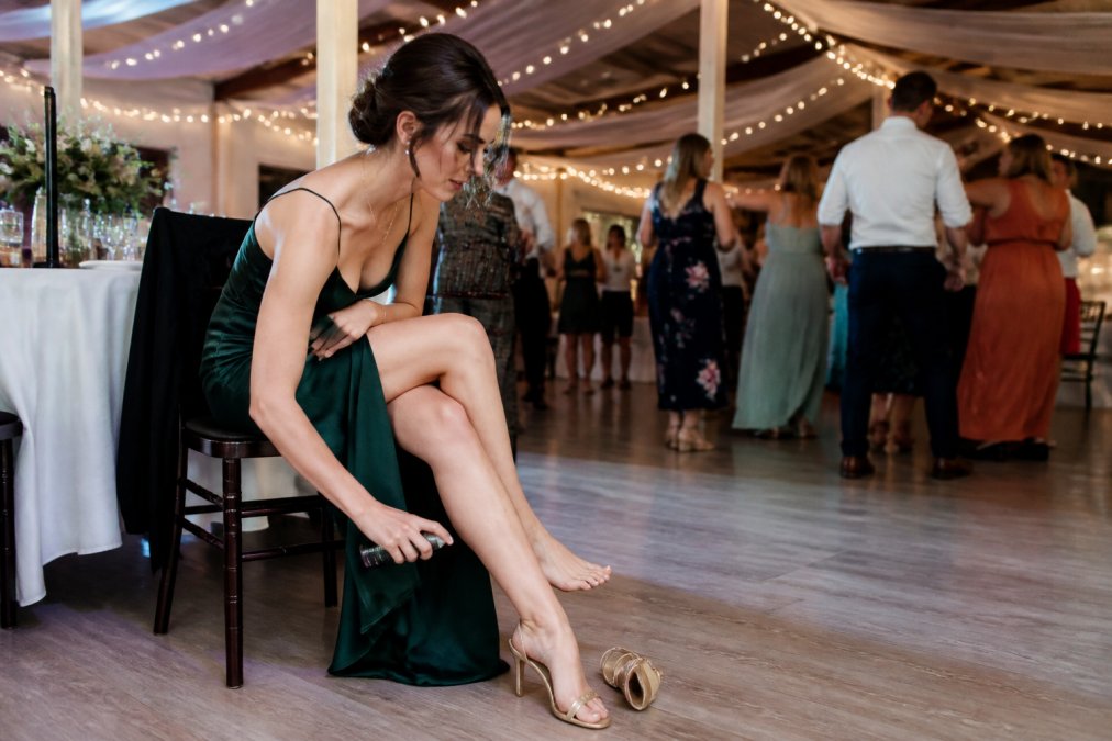 A woman in elegant high heels sitting at a wedding reception, discreetly spraying a small bottle on her feet under the table, with other guests dancing in the background
