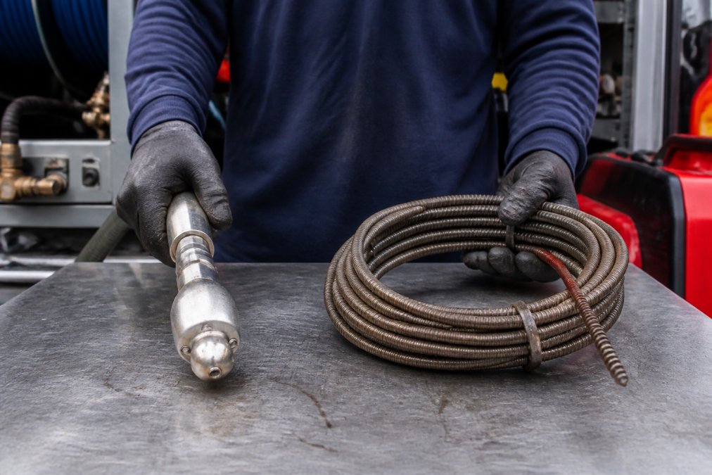 Close-up of a plumber's gloved hands holding a hydro jetting nozzle alongside a coiled drain snake cable, both laid out on a metal workbench inside a plumbing service van