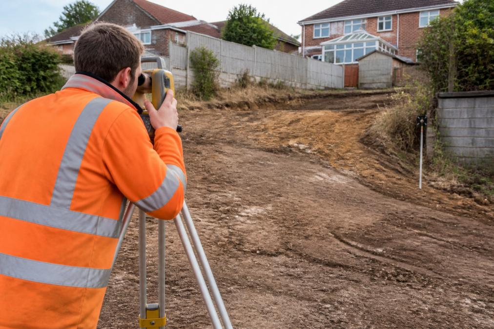 Professional surveyor using laser level on residential property to measure site elevation and drainage fall gradient, with slope direction marked on ground