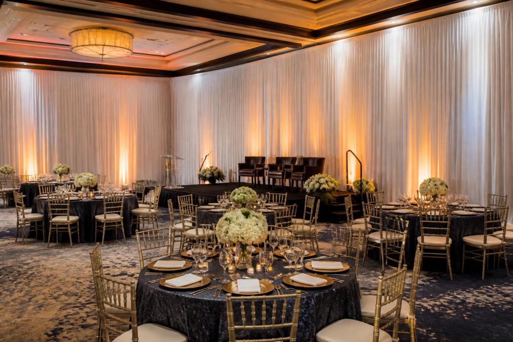 A banquet hall transformed with floor-to-ceiling white sheer drapes and a pipe and drape backdrop, warm uplighting on the fabric, round tables set for a corporate gala in the foreground