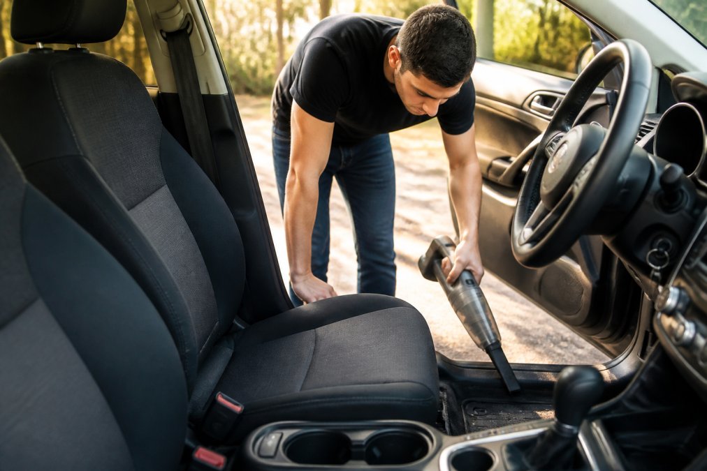 Person using cordless portable vacuum on car interior upholstery and floor mats with natural sunlight streaming through open door