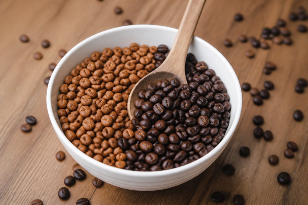 Close-up of whole coffee beans in a white ceramic cupping bowl with a wooden spoon, showing distinct color variation between light golden beans and darker chocolate-brown beans, with scattered beans on a natural wood surface
