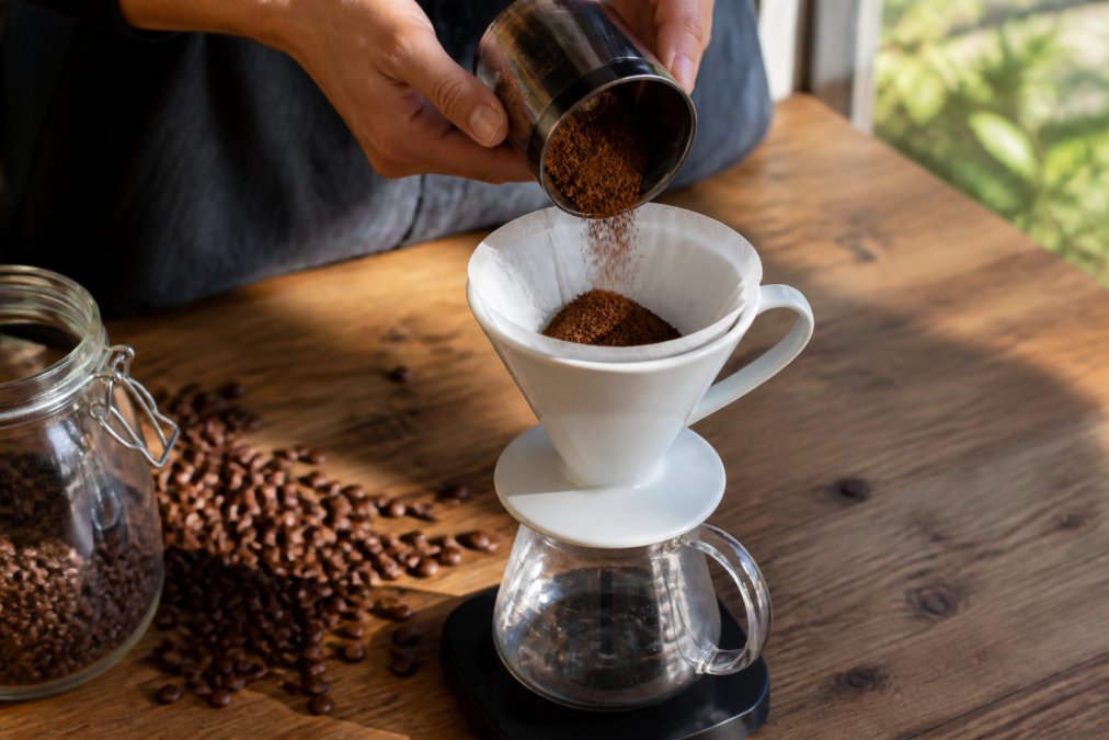 Hands carefully pouring freshly ground medium-roast coffee from a grinder into a white ceramic pour-over dripper, with whole beans scattered across a natural wooden table beside a glass carafe catching the morning sunlight
