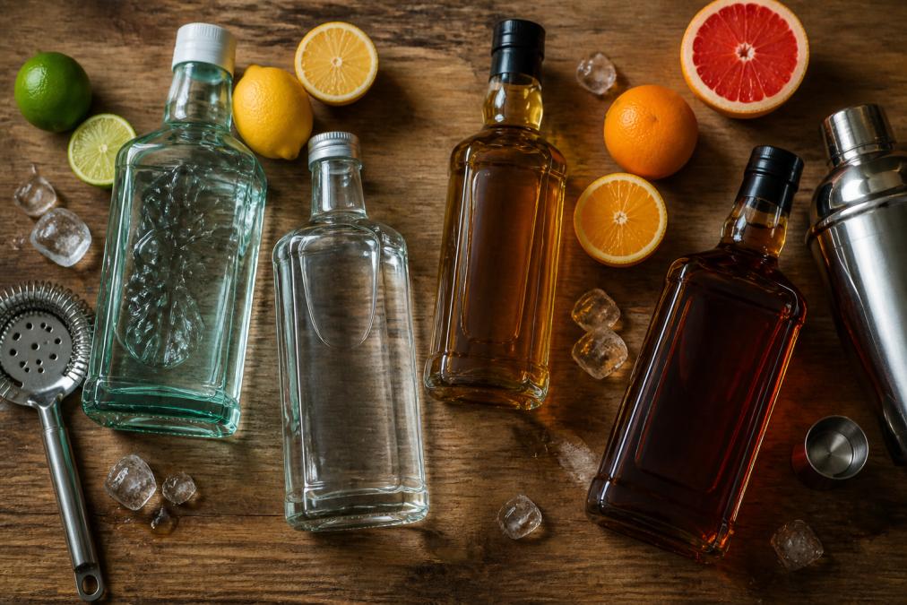 Overhead shot of gin, vodka, rum, and whiskey bottles arranged on a wooden bar counter with fresh citrus halves, ice cubes, and a metal shaker nearby