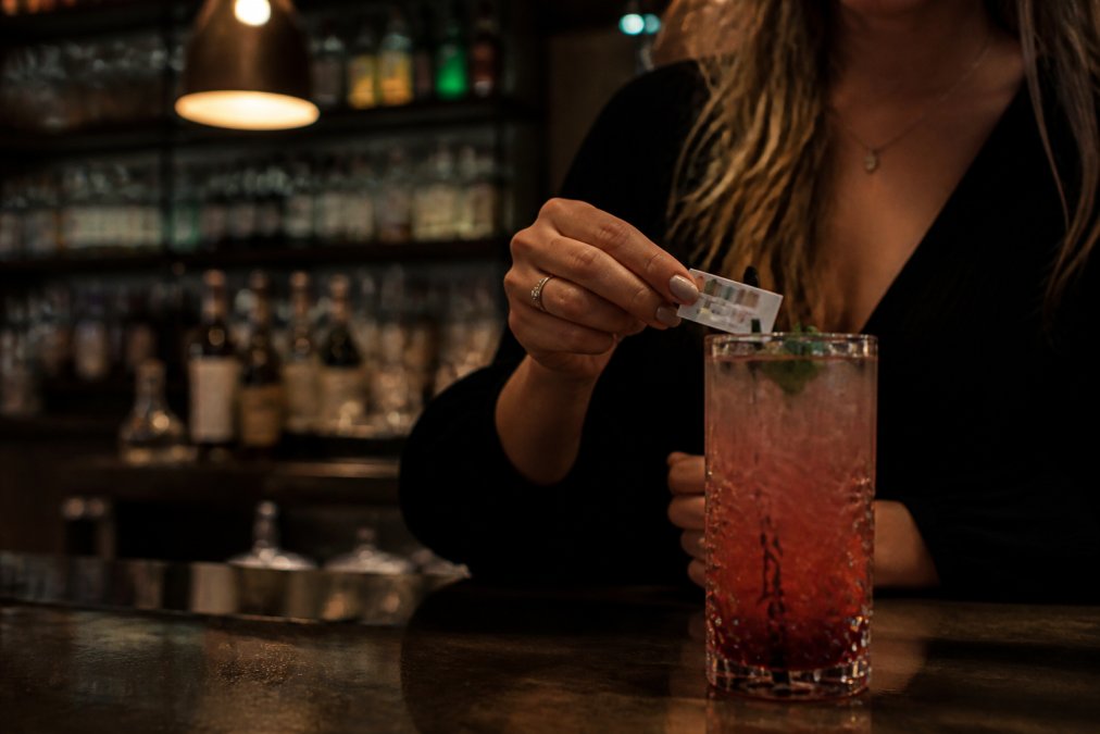 Close-up of a person discreetly placing a small test strip card on the edge of a drink glass at a dimly lit bar, with a colourful cocktail in the foreground and soft ambient lighting from bar fixtures overhead