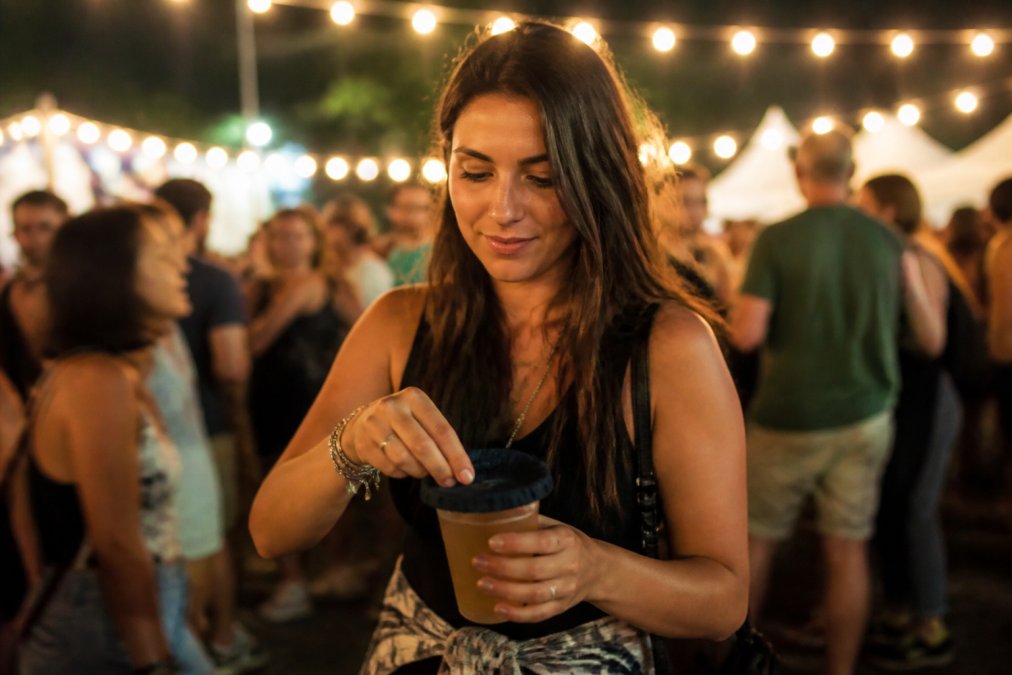 Young woman at an outdoor evening festival placing a fabric drink cover over her plastic cup, crowd visible in soft bokeh in the background, warm string lights overhead casting a golden glow