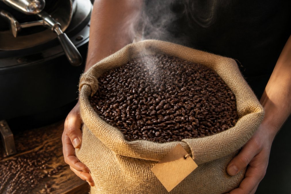 Close-up of freshly roasted coffee beans in a burlap bag with a roasted date label visible, steam rising from the beans, hands holding the bag in natural morning light