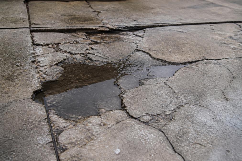 Close-up of a concrete driveway surface showing multiple cracks, spalling edges, and dark water pooling in depressions, with visible deterioration patterns spreading across the pavement