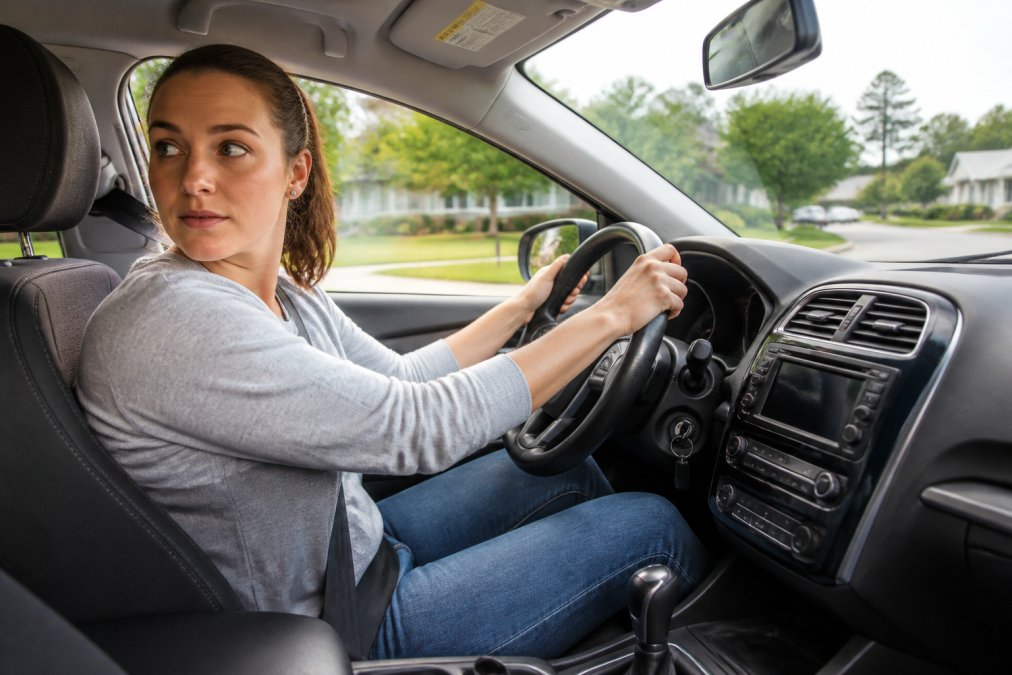 An adult learner gripping the steering wheel of a clearly marked driving school car, checking mirrors carefully while navigating a suburban street on a bright day, with focused expression and proper hand position