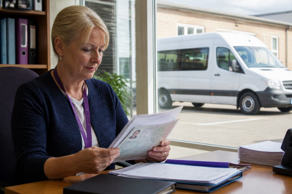 School business manager reviewing DVLA licence documentation at a desk with a minibus visible through the window in the background