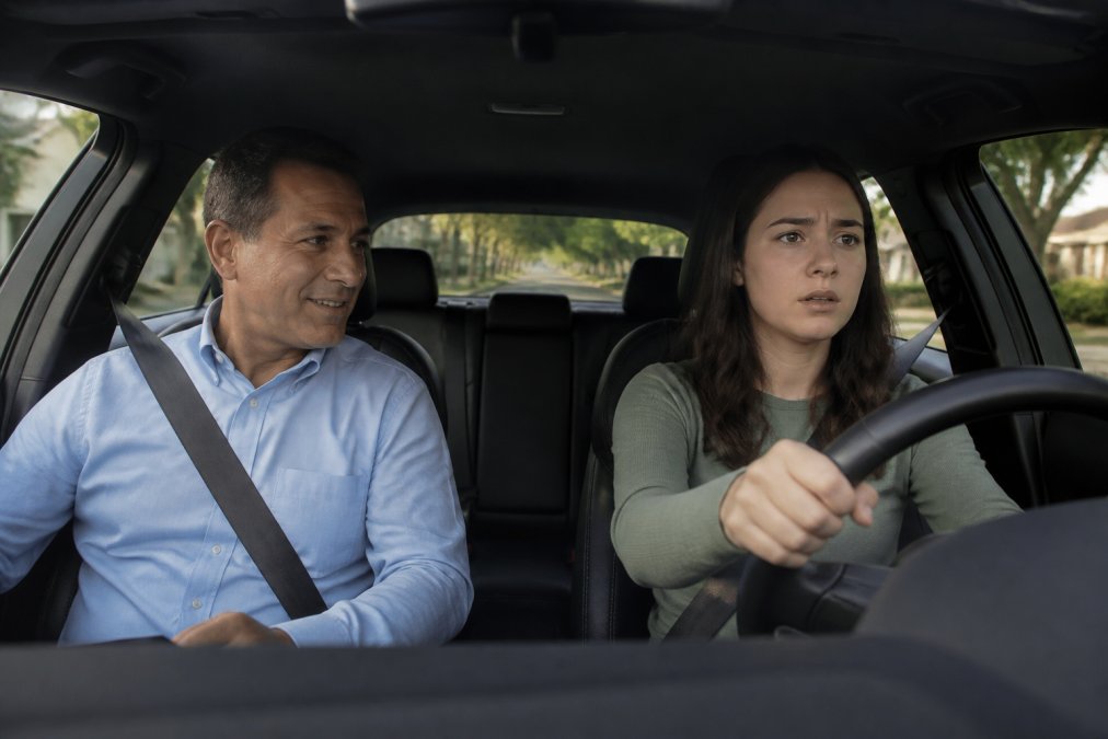 A calm, patient driving instructor sitting in the passenger seat of a dual-controlled training vehicle, smiling reassuringly at a nervous young adult driver with hands gripping the steering wheel, with a quiet residential street and tree-lined road ahead in soft afternoon light