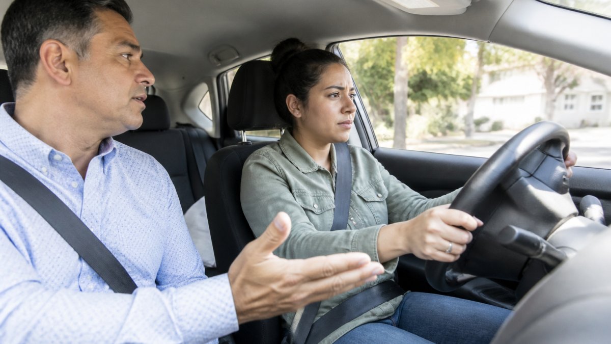 A calm, patient driving instructor sitting in the passenger seat, offering reassuring guidance to a nervous student driver during a behind-the-wheel lesson, both focused on the road ahead with natural daylight streaming through the windshield