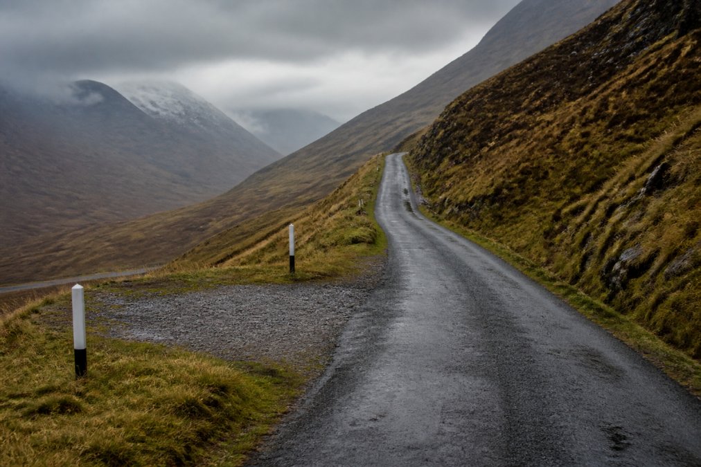 A narrow single-track Highland road stretching into misty mountains, a small passing place marked with white posts visible on the left verge, dramatic moorland rising on both sides under grey Scottish sky