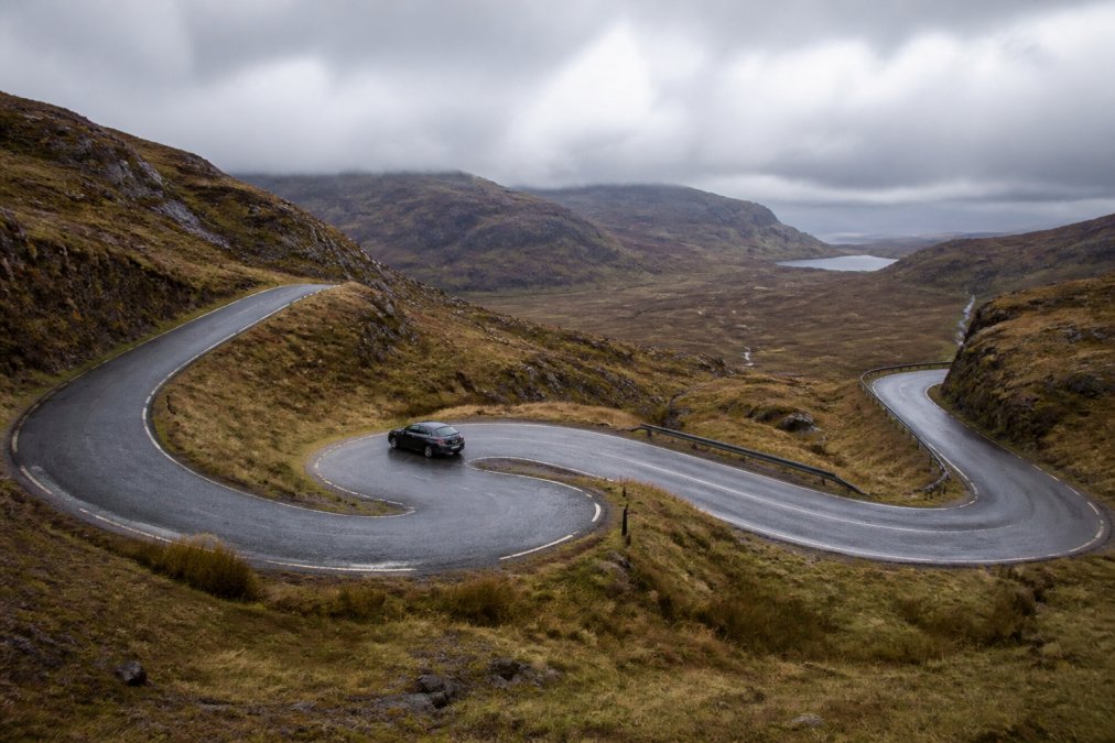 A winding Highland mountain pass with sharp hairpin bends, a small vehicle carefully navigating a tight curve, moorland slopes rising steeply on both sides, overcast sky with low cloud cover