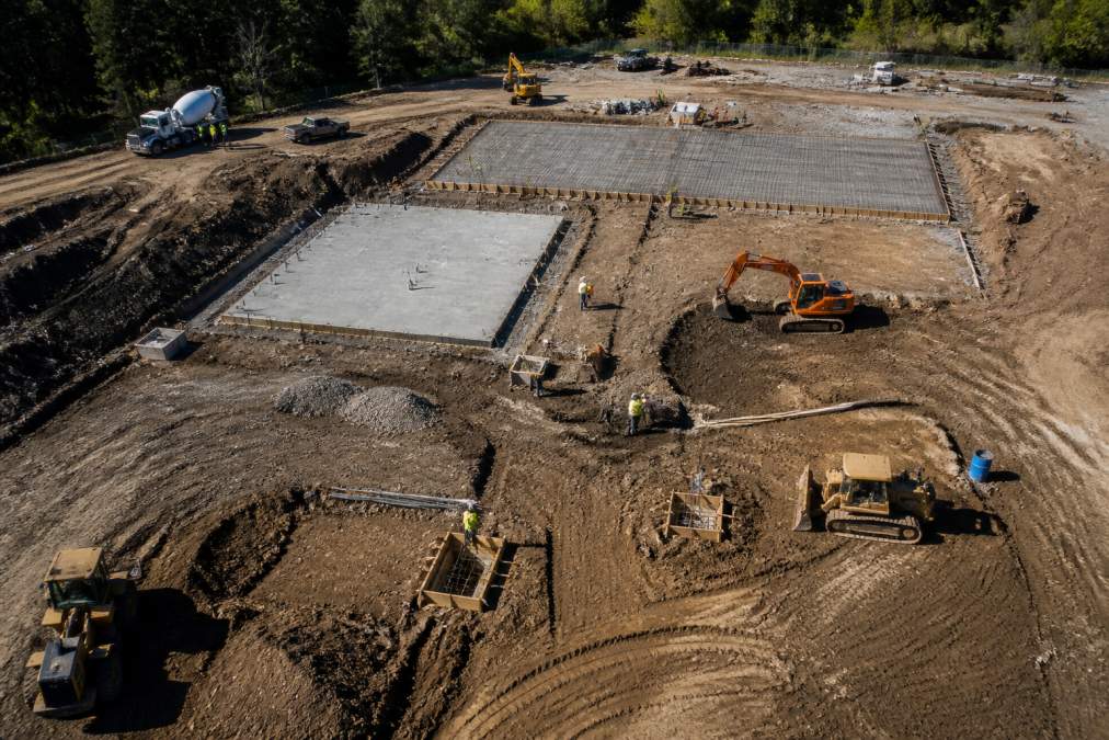 Aerial view of an active construction site from drone perspective showing multiple equipment, workers in safety gear, and progress markers visible from above with clear weather conditions