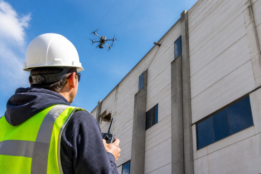 Certified drone pilot operating an industrial drone with advanced camera equipment during a building facade inspection, the drone positioned safely away from the structure with clear sky visible