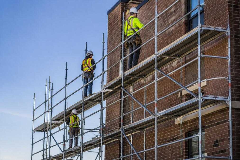 Metal scaffolding structure erected against a commercial building with safety railings, hard hats, and safety equipment visible on the platform, workers in high-visibility clothing