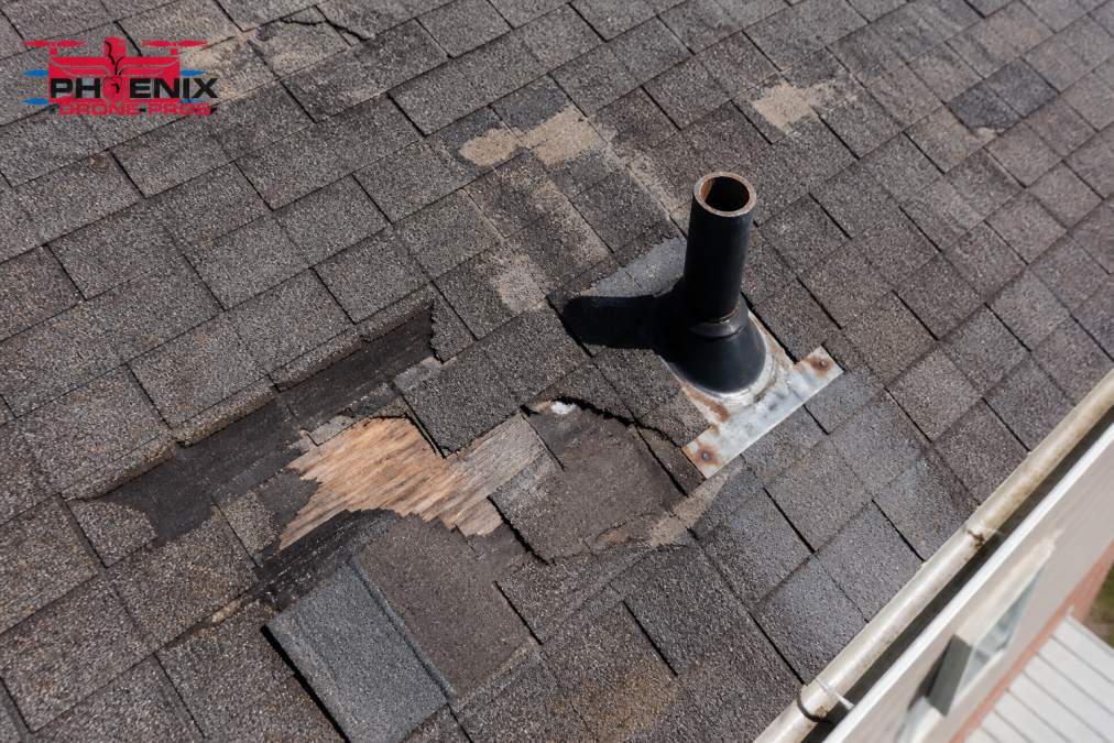 Close-up aerial view of a residential roof surface showing damaged and missing asphalt shingles, visible flashing issues around a vent pipe, and wear patterns visible from drone perspective hovering above