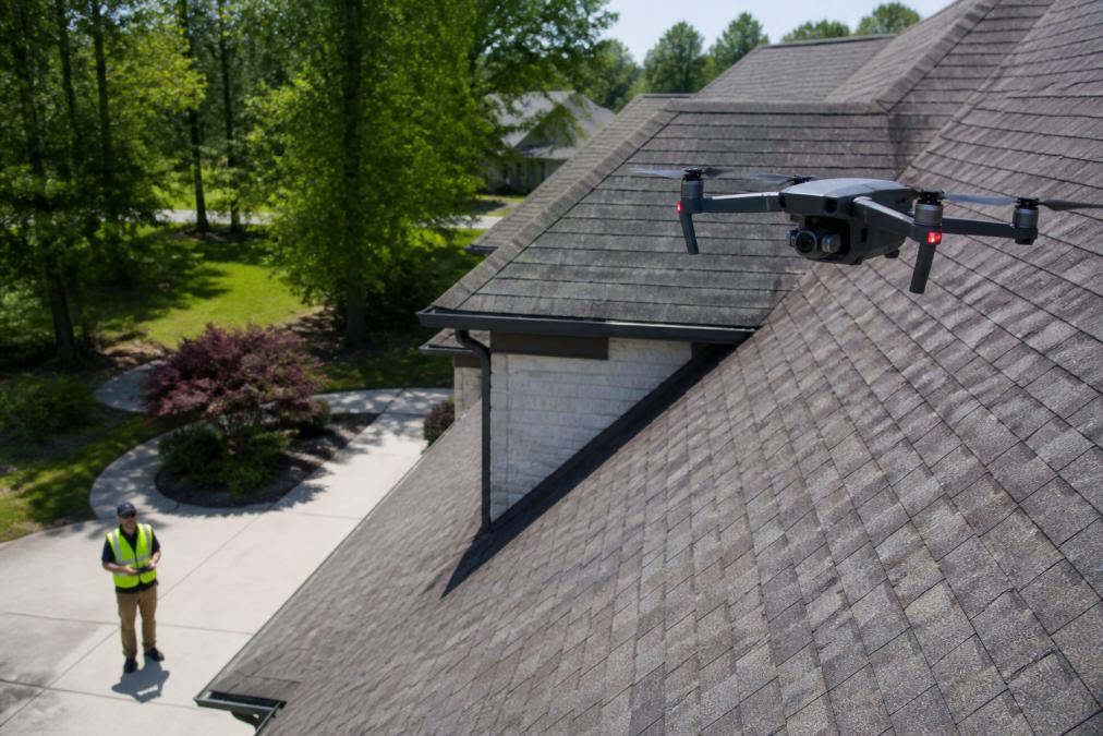 Commercial quadcopter drone hovering close to a steep residential roof in bright midday sunlight, capturing high-resolution footage while a ground crew member in a hi-vis safety vest observes from the driveway below