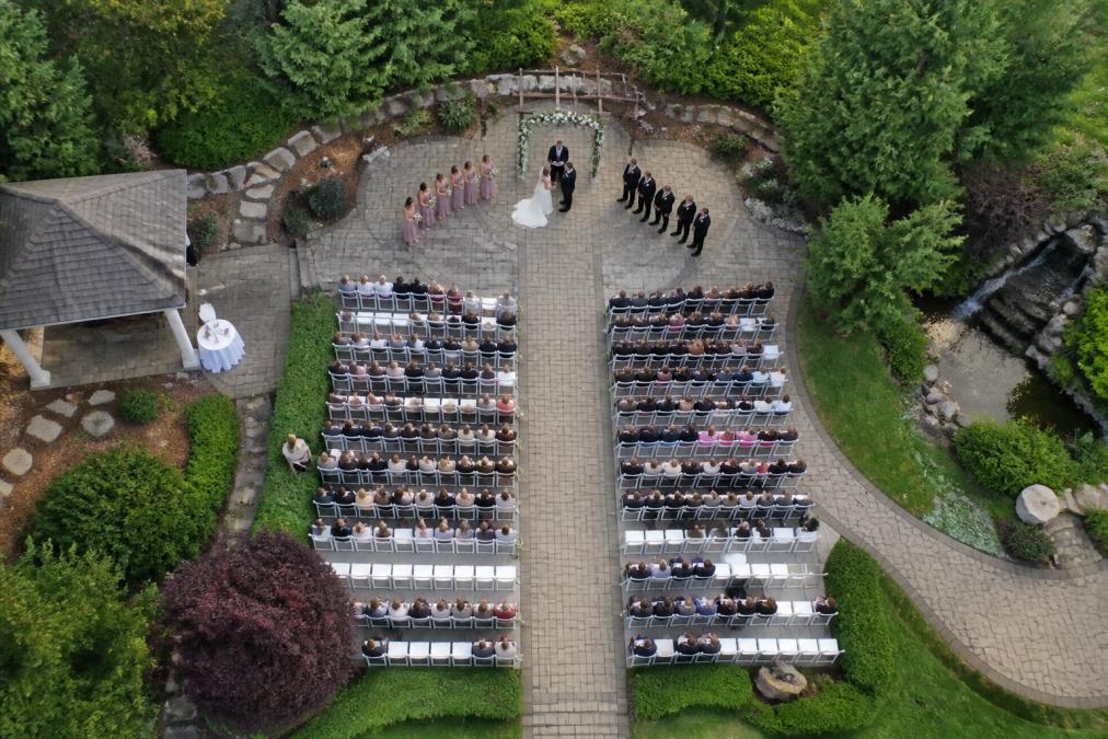 Aerial view of a wedding ceremony in progress, showing the bride and groom at the altar with guests seated on both sides in rows, captured from above at approximately 150 feet altitude, demonstrating the cinematic bird's-eye-view perspective and wide-angle composition that reveals the full venue layout and guest arrangement