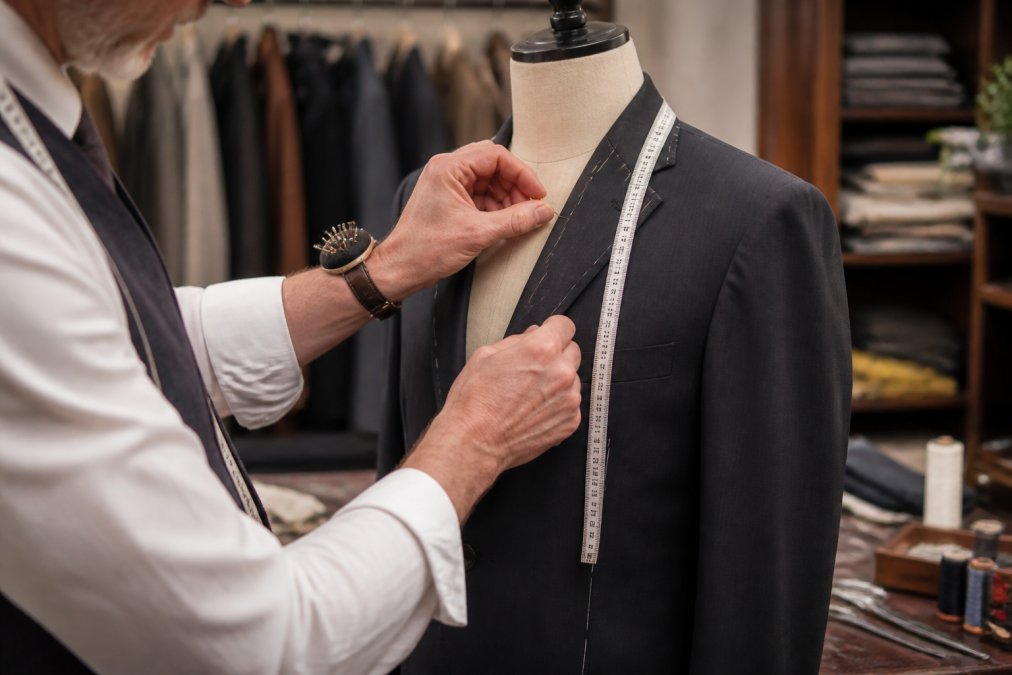 Close-up of tailor's hands pinning and measuring a suit jacket on a fitting form with precision tools visible, professional tailoring workstation in background
