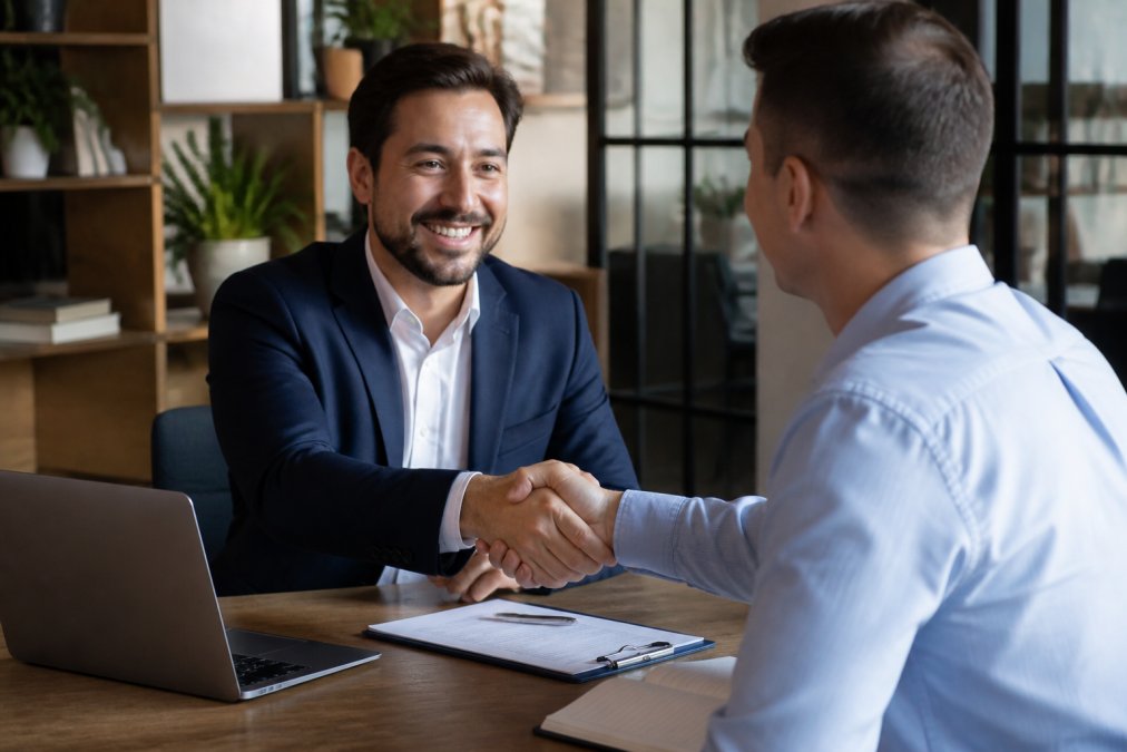 Professional loan officer and real estate investor shaking hands in a modern office after completing loan approval, with documents and laptop visible on desk