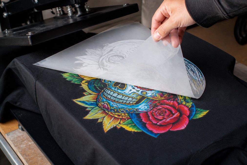 Close-up of a DTF transfer being heat-pressed onto a black cotton t-shirt on a production table, with the film partially peeled back to reveal a vibrant, full-color graphic underneath