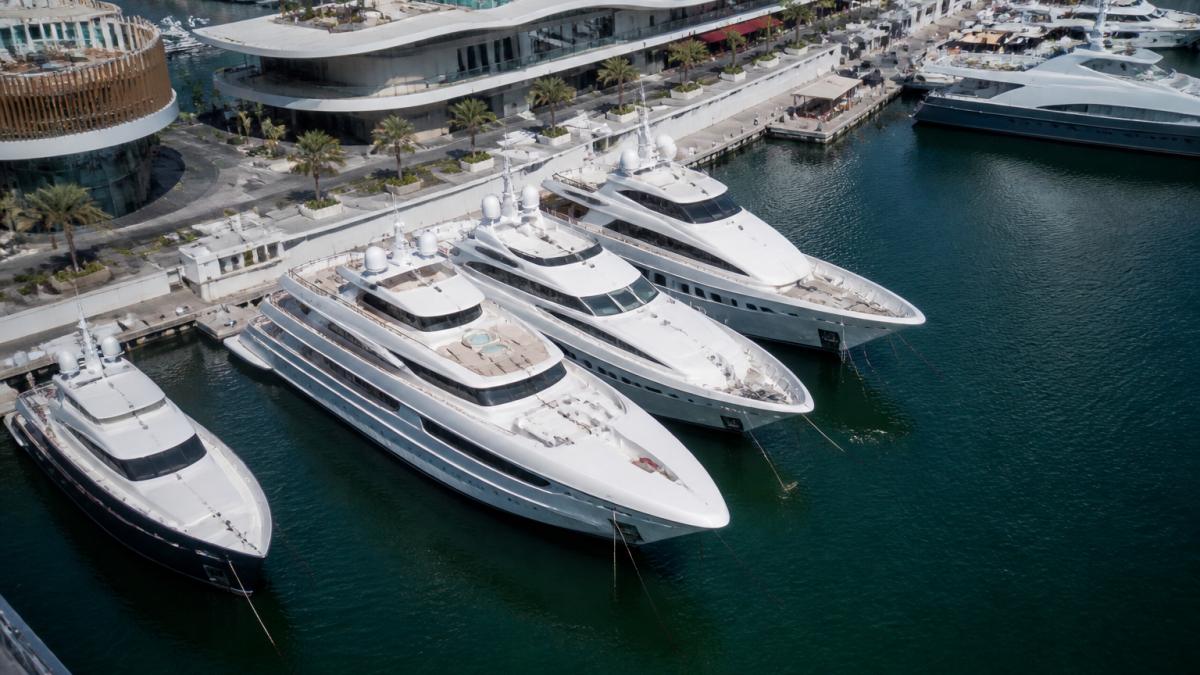 Aerial view of luxury superyachts lined up at Dubai Harbour during the boat show, with modern marina facilities, gleaming white hulls reflecting in calm water, and contemporary harbour architecture in the background