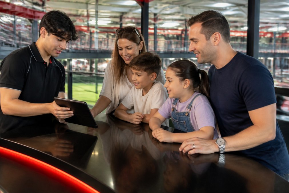 Family at a bright, modern karting venue reception desk, with a staff member in uniform showing booking details on a tablet computer while parents and excited children look on attentively