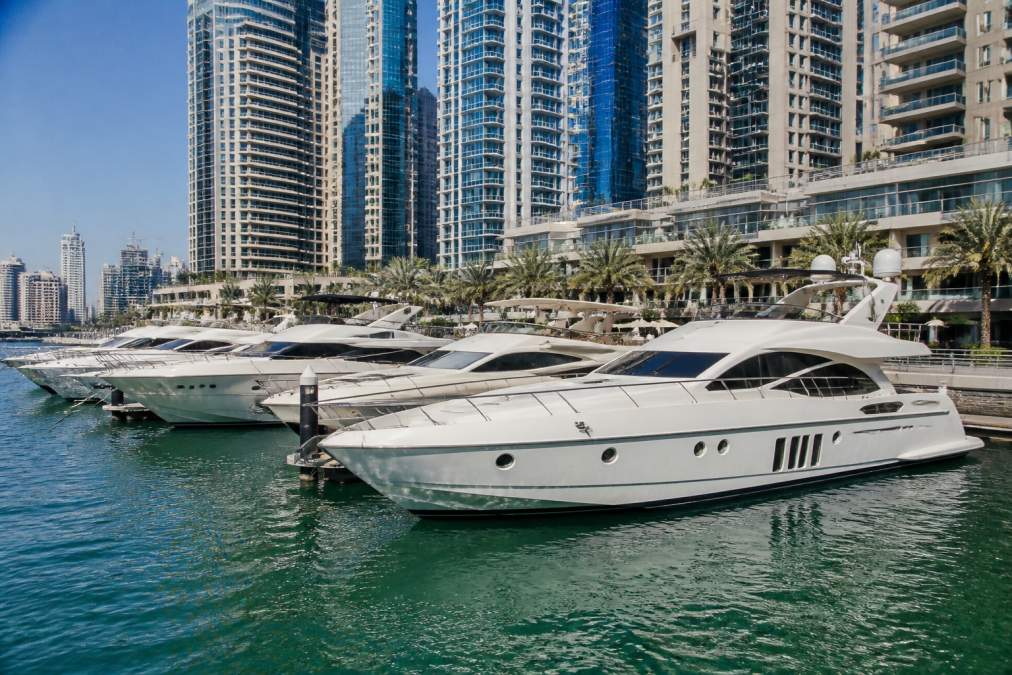 Luxury superyachts moored at Dubai Marina with modern high-rise buildings and waterfront promenade in background, daytime with clear blue water and white vessels