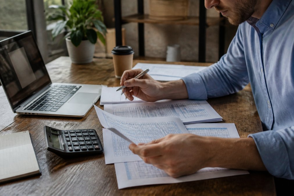 Business owner reviewing financial documents and spreadsheets at wooden desk with calculator, coffee cup, and laptop displaying charts in natural office lighting