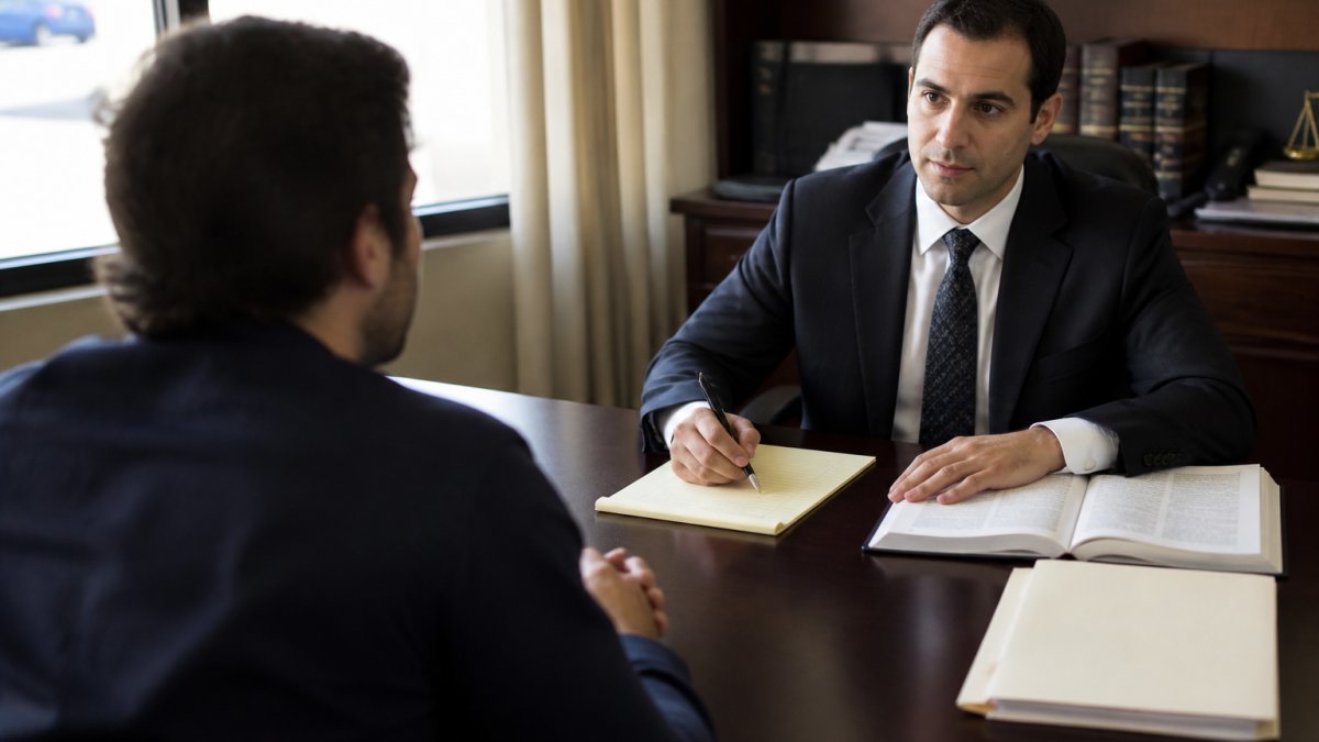 Professional attorney in business attire sitting at desk across from client during consultation, with legal documents and notepad visible, natural office lighting