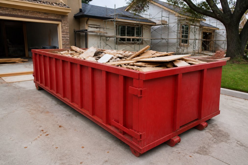 A large roll-off dumpster positioned in a residential driveway during an active home renovation project, with construction materials and debris visible inside the container