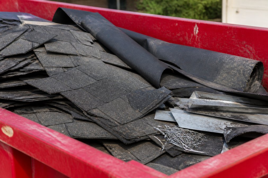 Close-up of asphalt shingles, felt underlayment, flashing pieces, and roofing nails stacked together in natural daylight