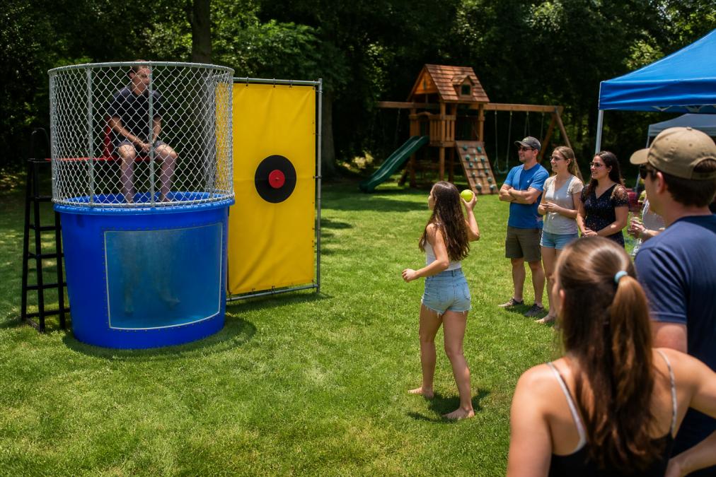 A clean, modern dunk tank rental set up on a grassy lawn at a sunny outdoor party, with a safety ladder and clear water visible, guests gathered around laughing