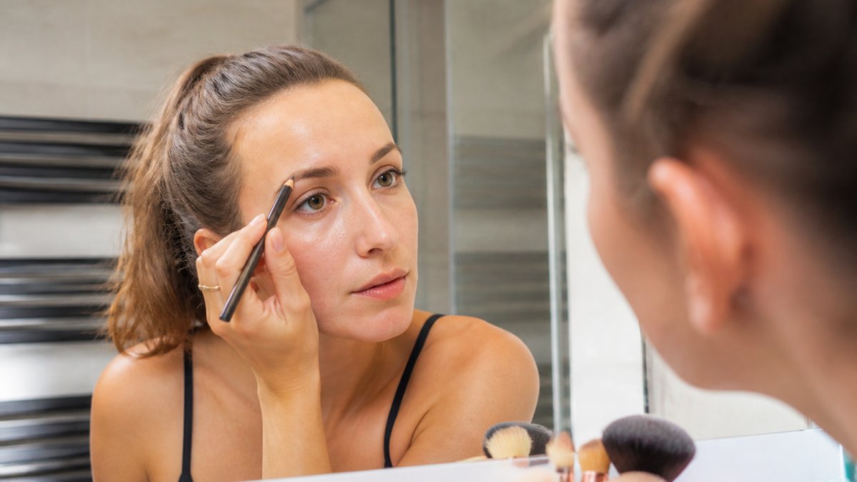 Vrouw gebruikt een wenkbrauwpotlood om dunne wenkbrauwen bij te werken voor de spiegel in een heldere badkamer