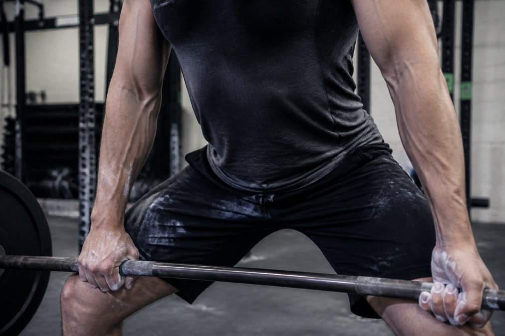 Close-up of an athlete's chalked hands gripping a barbell during a heavy deadlift, showing the friction point where the bar contacts their training top and forearms in a gritty gym setting