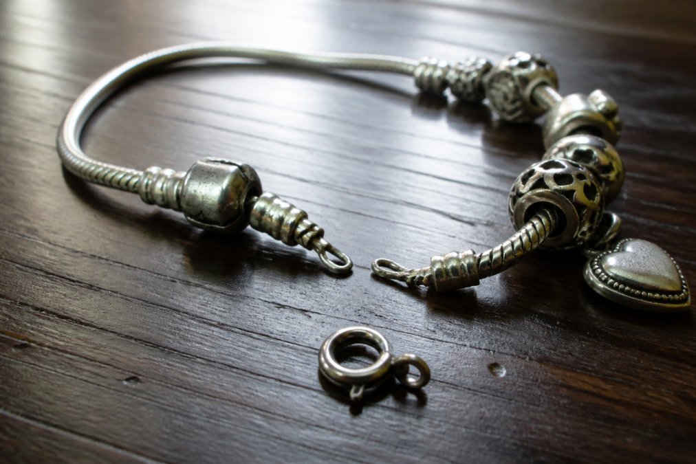 Close-up of a worn silver charm bracelet with a bent link and a broken spring-ring clasp lying on a dark wooden table, soft window light revealing scratches on the metal surface