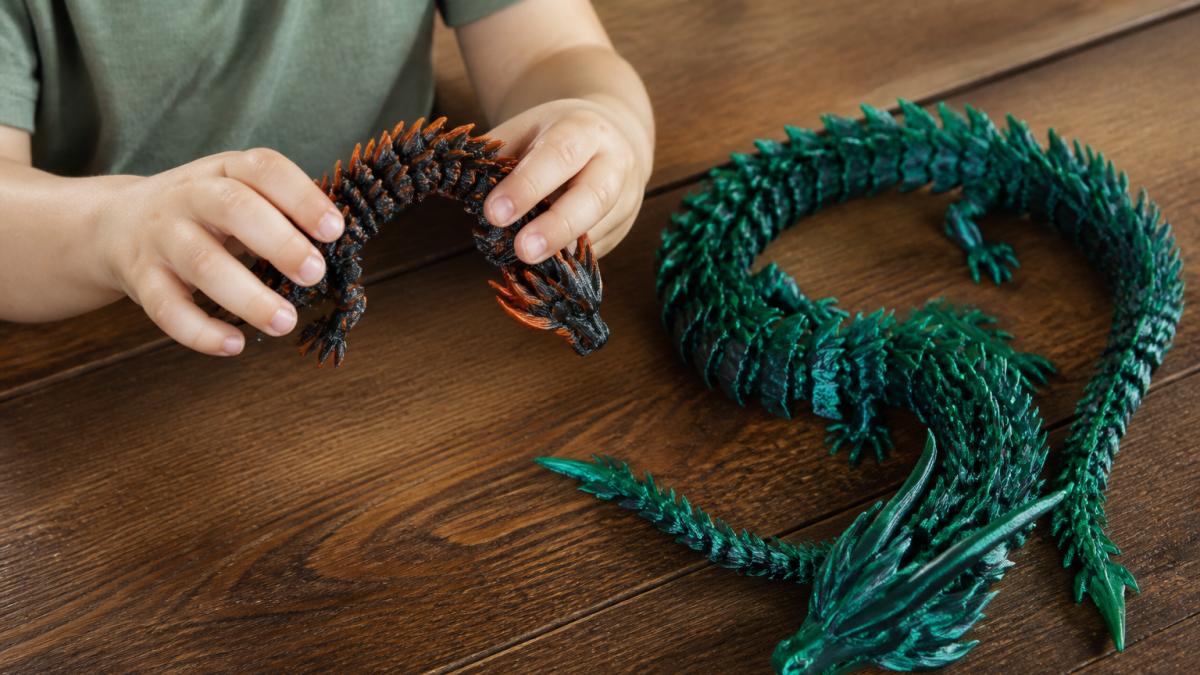 A child's hands bending the flexible segmented body of a 3D-printed articulated dragon on a wooden table, with a second larger dragon coiled nearby for scale