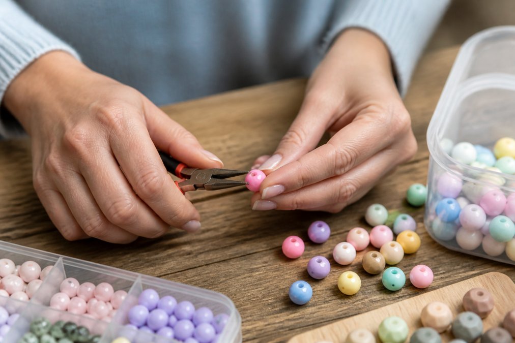A crafter's hands holding a silicone bead and a pair of pliers, testing its flexibility on a wooden workbench with other beads scattered around