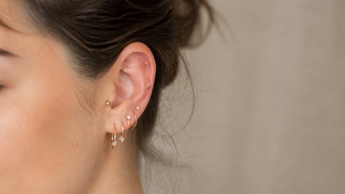 Close-up of a woman with multiple ear piercings wearing small gold and titanium studs and hoops, soft natural lighting, neutral background, showing healed piercings and delicate jewelry
