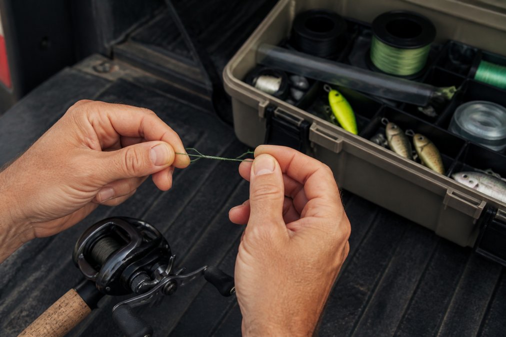 Angler's hands demonstrating proper knot tying technique with braided fishing line near a baitcasting reel and open tackle box