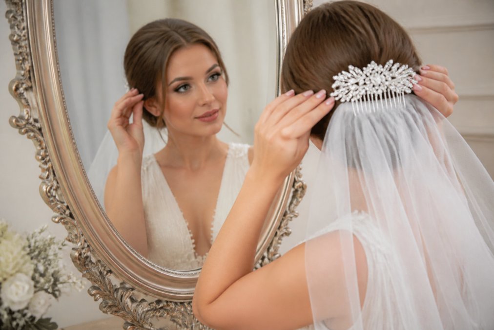 Bride adjusting her delicate silver wedding veil and crystal hair comb while looking in an ornate mirror, wearing a classic ivory gown with soft tulle layers
