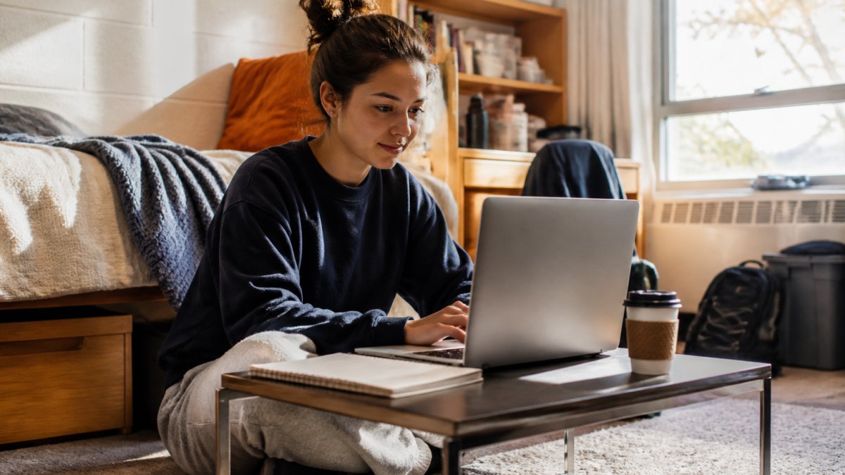 A student sitting cross-legged on a dorm room floor with a slim laptop open on a low table, notebook and coffee cup beside it, warm afternoon light through a window